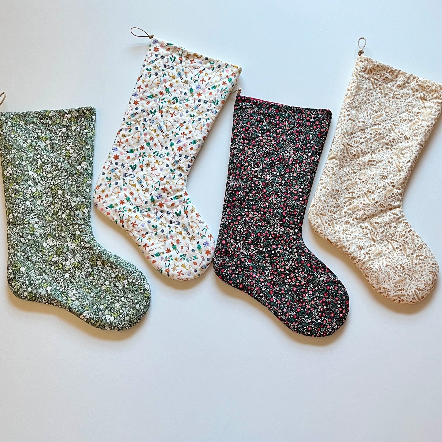 Four Christmas stockings in various colors and patterns, including a green plaid, a green and red stripe, a blue stripe, and a pine forest design, all quilted and lined with a solid color fabric.