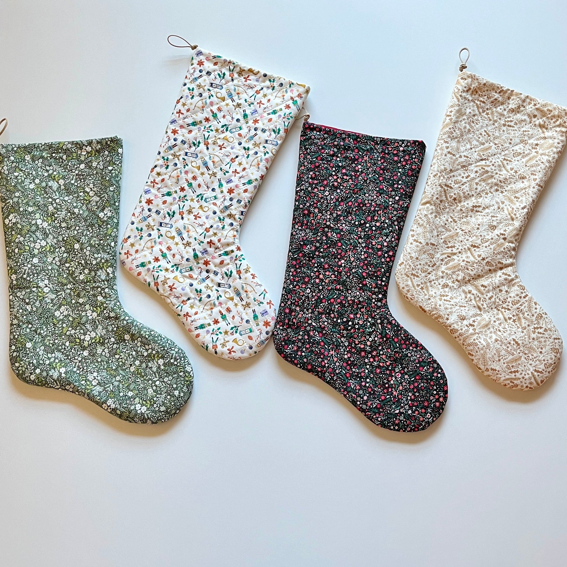 Four Christmas stockings in various colors and patterns, including a green plaid, a green and red stripe, a blue stripe, and a pine forest design, all quilted and lined with a solid color fabric.